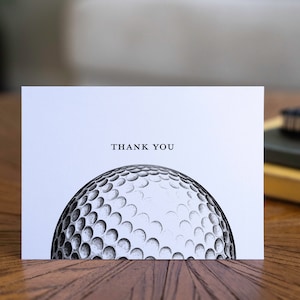 Könnte beinhalten: Eine weiße Dankeskarte mit dem schwarzen Aufdruck "THANK YOU". Die Karte zeigt eine detaillierte Illustration eines Golfballs. Die Karte liegt auf einer Holzoberfläche.