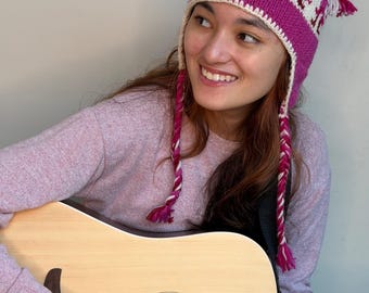 May include: A knit beanie hat featuring a vibrant magenta and cream color scheme with a repeating geometric pattern. The hat includes ear flaps and two long, braided tassels in magenta and cream that hang down from the sides of the head.