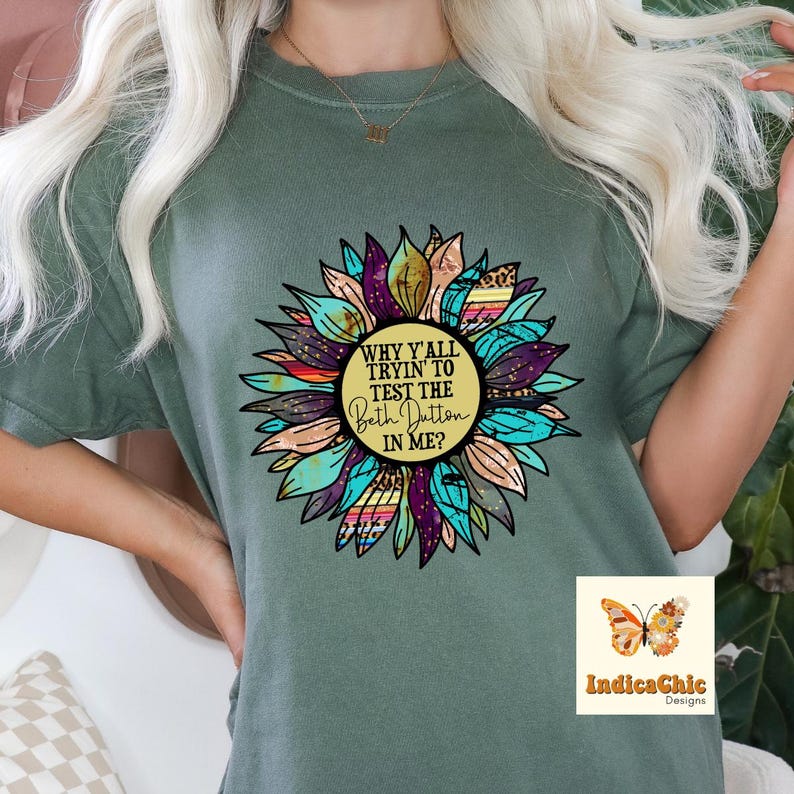 May include: A green t-shirt with a colorful sunflower design. The sunflower has a yellow center with the text "Why y'all tryin' to test the Beth Dutton in me?"  The sunflower has a variety of colors including turquoise, purple, orange, and brown.