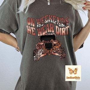 K&ouml;nnte beinhalten: Ein graues T-Shirt mit dem Text "On Weekends We Wear Dirt" in einer braunen und Leopardenmuster-Schriftart. Unter dem Text befindet sich ein braunes ATV mit Leopardenmuster. Der Text "IndicaChic Designs" befindet sich in der rechten unteren Ecke des Bildes.