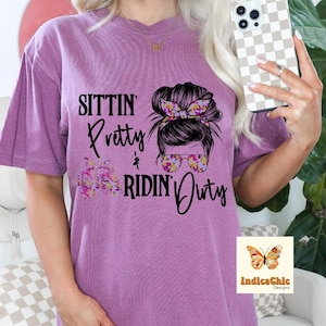 Puede incluir: Camiseta morada con un gráfico de una mujer con un moño desordenado y gafas de sol. El texto "Sittin' Pretty & Ridin' Dirty" está impreso en la camiseta.