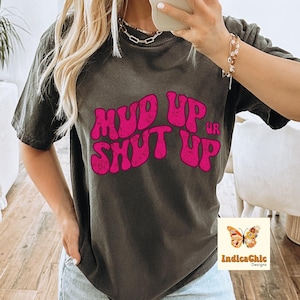 以下が含まれることがあります： 濃いグレーのTシャツに、レトロなフォントで「Mud Up or Shut Up」と書かれたピンクのグラフィックがプリントされています。