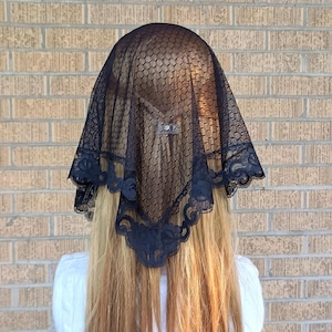 May include: A black lace veil with a scalloped edge and floral details. The veil is draped over a person's head, with long blonde hair visible. The person is wearing a white sweater. The background is a brick wall.