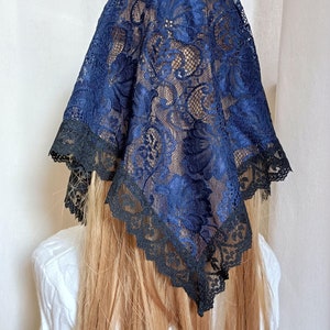 May include: A navy blue lace head covering with a floral design and a black scalloped edge. The head covering is draped over a person's head and shoulders, with long blonde hair visible.