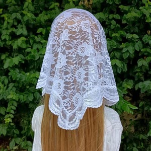May include: A white lace head covering featuring a detailed floral pattern with scalloped edges. The fabric is semi-sheer and delicate, draped over the head to form a triangular shape that rests against the back, showcasing intricate needlework.