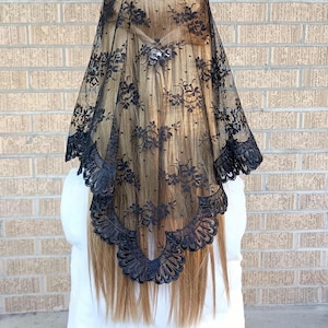 May include: A black lace veil featuring a detailed floral pattern throughout the fabric. The veil has a scalloped edge with a decorative border design. It is draped over long, light brown hair, covering the back of the head and shoulders.
