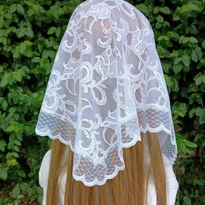 May include: A white lace chapel veil featuring a delicate floral pattern with scalloped edges. The sheer fabric is draped over long, light brown hair, showcasing the intricate embroidery and the fine mesh texture of the triangular head covering.