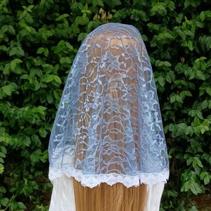 May include: A light blue lace head covering featuring a delicate floral pattern and a scalloped white lace trim along the bottom edge. The fabric is semi-sheer, allowing the texture of the lace to be clearly visible against the light blue base.