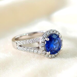 Könnte beinhalten: Ein silberner Ring mit einem großen, runden, tiefblauen Edelstein in der Mitte, umgeben von einem Kranz aus kleineren, klaren Steinen. Das Band teilt sich in zwei Reihen kleinerer Steine auf beiden Seiten des Hauptsteins auf.