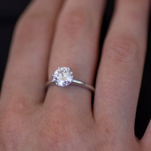 May include: A silver engagement ring with a large, round, clear gemstone. The ring is set on a thin band and is displayed on a person's finger. The background is dark.