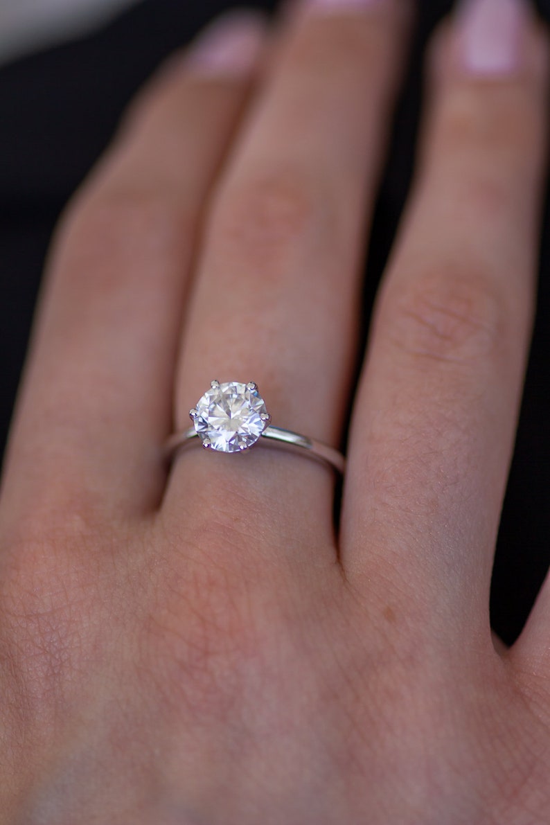 May include: A silver engagement ring with a large, round, clear gemstone. The ring is set on a thin band and is displayed on a person's finger. The background is dark.