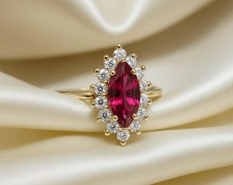 Op de afbeelding: Een gouden ring met een marquise-geslepen robijn, omringd door kleine, ronde diamanten. De ring heeft een slanke, gepolijste band, tegen een zachte, crèmekleurige achtergrond.