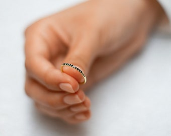 May include: A delicate gold ring with a row of small, black gemstones. The ring is held in a hand, showcasing its slender design and the contrast between the gold band and the dark stones. The ring is a piece of jewelry.