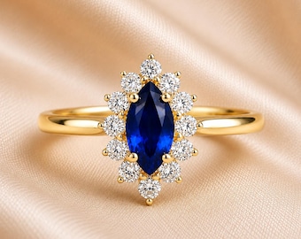 May include: A gold ring featuring a central marquise-cut deep blue gemstone surrounded by a halo of twelve small round clear stones. The band is smooth and polished gold, creating a bright contrast against the dark blue centre stone.