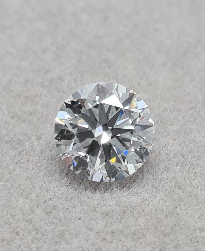 May include: A round-cut diamond with numerous facets, reflecting light. The colorless gemstone is set against a light gray background. The diamond's brilliance is highlighted by the intricate cuts.