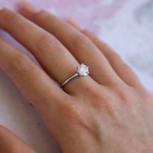 May include: A silver engagement ring with a large, round, clear gemstone. The ring is set on a slender band and is displayed on a person's hand with a soft pink background.