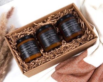 May include: A gift box filled with three amber glass candles. The candles are labeled "Teakwood Trails", "Twilight Forest", and "Pinewood Trails". Each candle has a black label with white text.