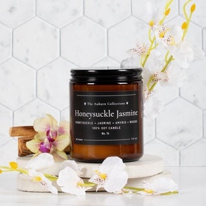 May include: A brown glass jar candle with a black lid, labeled "Honeysuckle Jasmine." The candle sits on a white stone with decorative flowers. The background is a white hexagon tile wall. The candle is 3.5 oz.