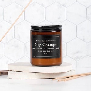 May include: A Nag Champa scented candle in a brown glass jar with a black lid. The label reads "The Auburn Collections" and lists the scent notes. The candle is 7.0 oz and made of 100% soy wax. It sits on a white, round platform.