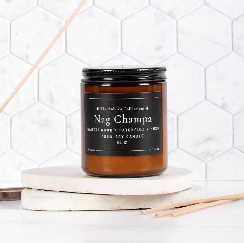 May include: A Nag Champa scented candle in a brown glass jar with a black lid. The label reads "The Auburn Collections" and lists the scent notes. The candle is 7.0 oz and made of 100% soy wax. It sits on a white, round platform.