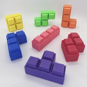 Peut inclure: Plusieurs pièces de puzzle géométriques colorées de différentes formes et couleurs, dont le jaune, le vert, l'orange, le bleu, le rose, le rouge et le violet. Les pièces sont disposées sur une surface blanche, rappelant le jeu Tetris classique.