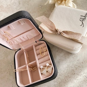 May include: Two small black and white jewelry boxes with a zipper closure. The black box is open and contains a gold chain necklace, a gold ring, and a pair of pearl earrings. The white box has a gold bow and the name "Abbie" written on it.