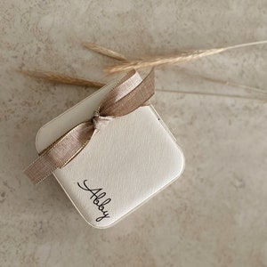 May include: A small, white, square box with a brown ribbon tied around it. The box has the word "Allie" written on it in black cursive.