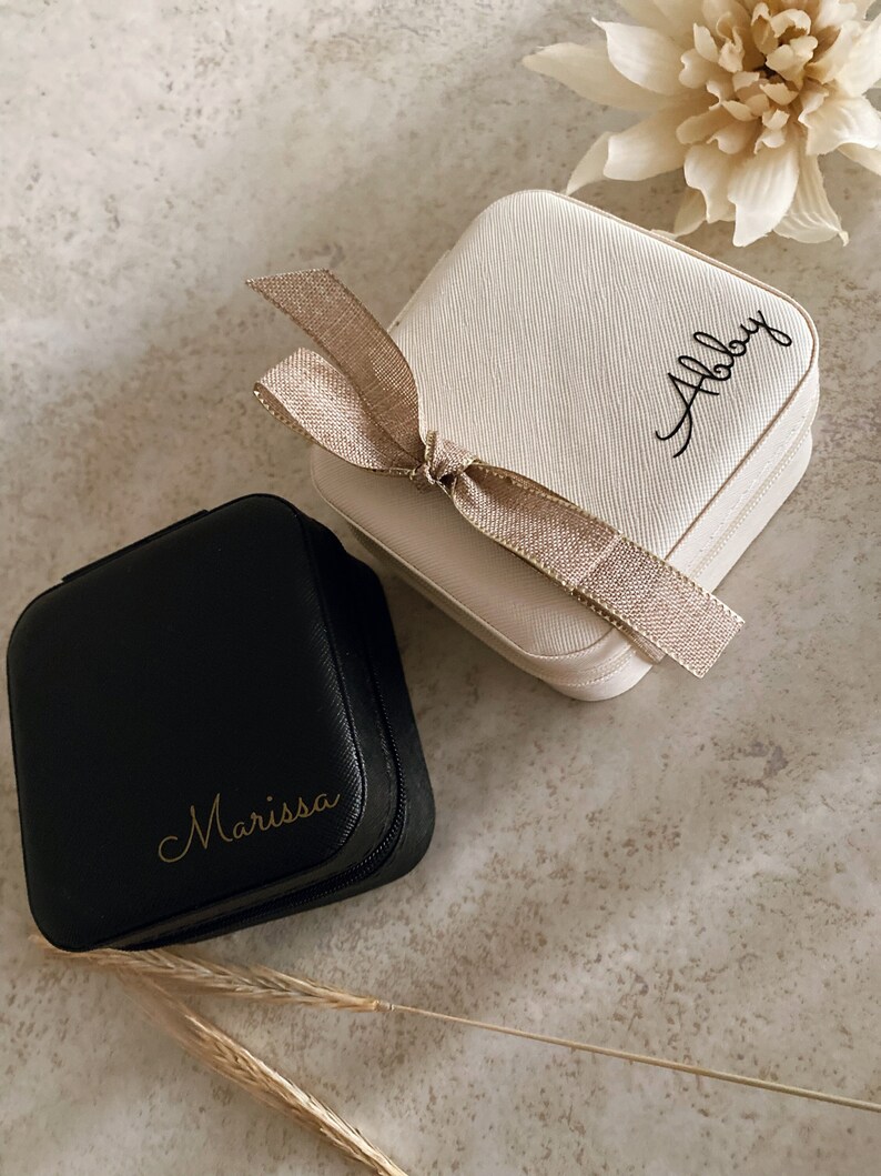 May include: Two small square jewelry boxes with zipper closures. The box on the left is black with the name "Marissa" written in gold lettering. The box on the right is white with a brown ribbon and the name "Abby" written in black lettering.