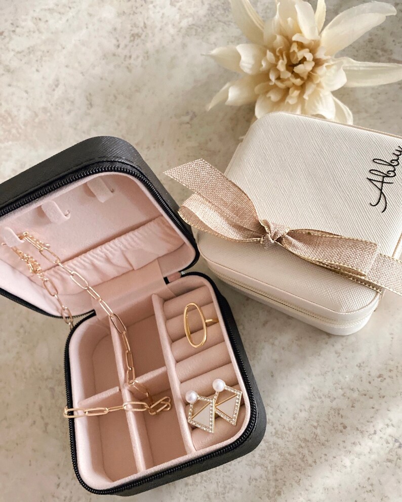 May include: Two small black and white jewelry boxes with a zipper closure. The black box is open and contains a gold chain necklace, a gold ring, and a pair of pearl earrings. The white box has a gold bow and the name "Abbie" written on it.