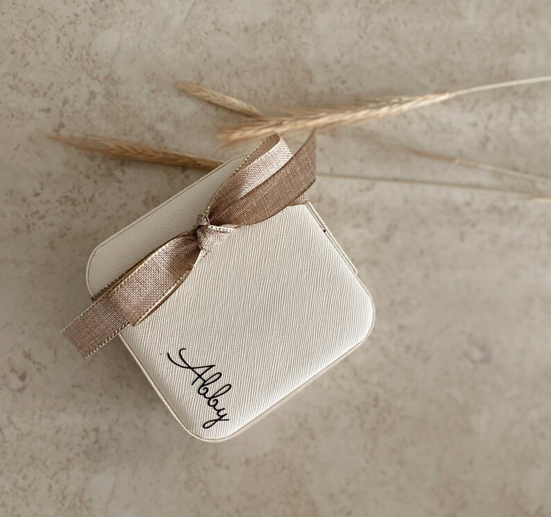 May include: A small, white, square box with a brown ribbon tied around it. The box has the word "Allie" written on it in black cursive.