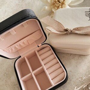 May include: Two small black and cream colored jewelry boxes with zipper closures. The black box is open and has a pink velvet interior with compartments for rings and earrings. The cream box has a gold ribbon and a gold zipper.