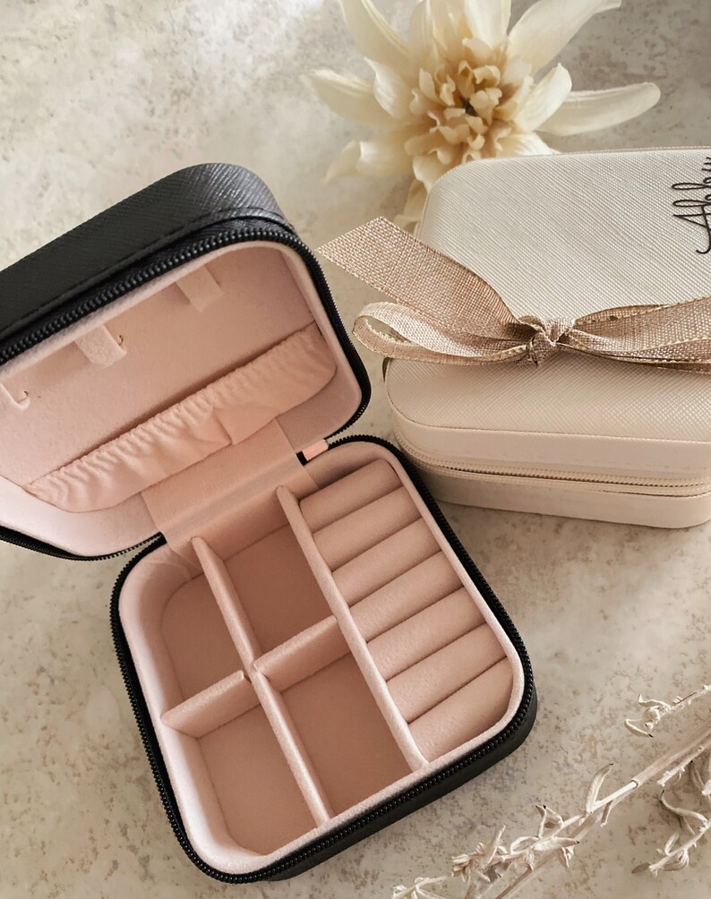 May include: Two small black and cream colored jewelry boxes with zipper closures. The black box is open and has a pink velvet interior with compartments for rings and earrings. The cream box has a gold ribbon and a gold zipper.