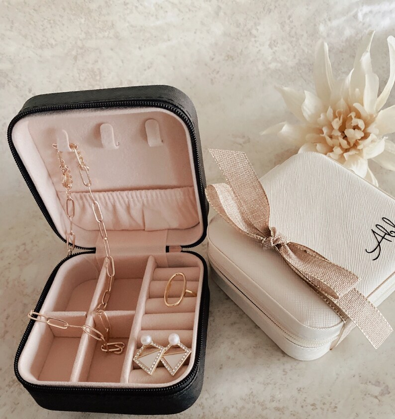 May include: Two small jewelry boxes, one black and one white. The black box is open and has compartments for rings, earrings, and necklaces. The white box has a gold ribbon and the name "Abby" written on it.