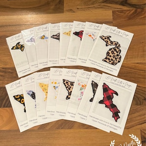 May include: A collection of white cards featuring embroidered patches in the shape of the Upper Peninsula of Michigan. The patches display various patterns, including sunflowers, bees, and plaid. The cards are arranged on a wooden surface.
