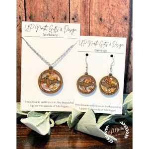 May include: A set of jewelry featuring a necklace and earrings with a wooden pendant and earrings shaped like the Upper Peninsula of Michigan. The pendant and earrings are decorated with gold glitter and a white background. The text "UP North Gifts & Design" is on the cards.