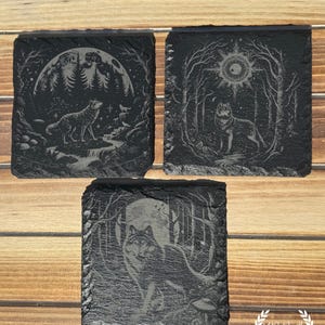 May include: Three square black slate coasters with etched wolf designs. One coaster features a wolf howling at the moon, another shows a wolf in a forest with a sun, and the third depicts a wolf in a forest with a full moon.