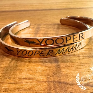 May include: Two gold-coloured metal cuff bracelets. One bracelet is engraved with the word "YOOPER" and a silhouette of a map. The other bracelet says "YOOPER MAMA". The bracelets are on a wooden surface.