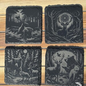 May include: Set of four square slate coasters with etched forest and wildlife designs. Each coaster features a different scene: a deer in a forest, a deer with a celestial design, a bear, wolf, and deer, and a deer, bear, and wolf under a moon.