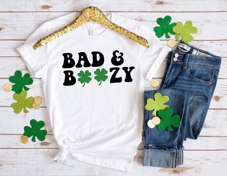 May include: White t-shirt with black text that reads "Bad & Boozy" with three green shamrocks. The shirt is on a white wooden background with green shamrocks and gold coins.