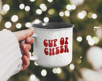 Pode incluir: Caneca de cerâmica branca com borda preta e um gráfico vermelho "Cup of Cheer" em uma fonte retrô. A caneca é salpicada de manchas pretas e está sendo segurada por uma mão.