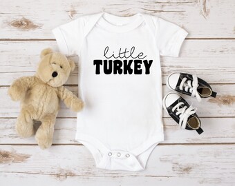 Puede incluir: Body de bebé blanco con el texto negro "little TURKEY". Un oso de peluche beige y un par de zapatillas de bebé negras y blancas están al lado del body. El body tiene mangas cortas y corchetes en la parte inferior.