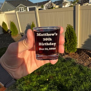 May include: A clear shot glass filled with a dark liquid, engraved with "Matthew's 30th Birthday Dec 21, 2026." The glass is held in a hand, with a blurred background of a yard, fence, and houses.