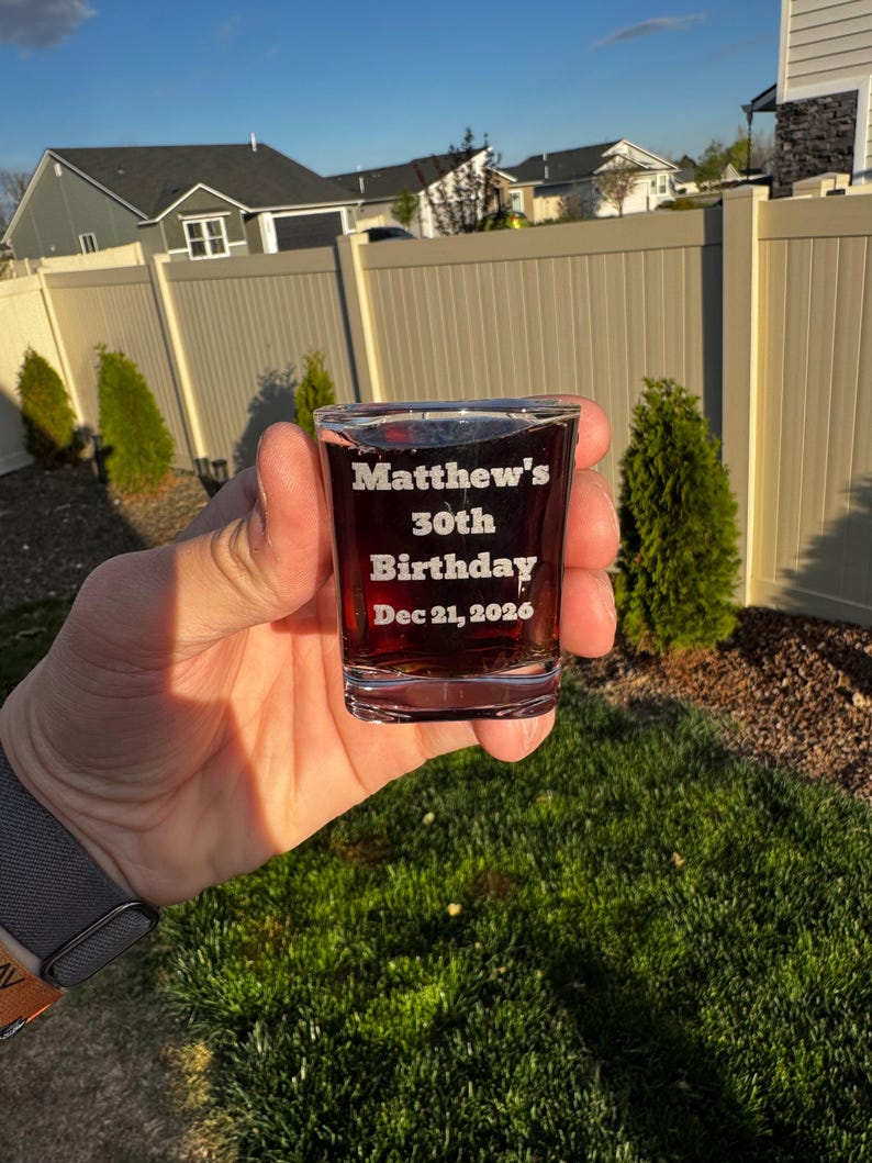 May include: A clear shot glass filled with a dark liquid, engraved with "Matthew's 30th Birthday Dec 21, 2026." The glass is held in a hand, with a blurred background of a yard, fence, and houses.