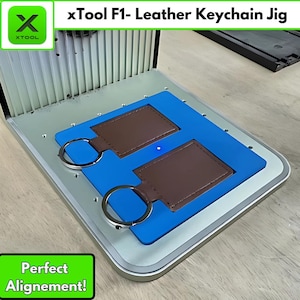 May include: An xTool F1 leather keychain jig with two brown leather keychains and silver key rings. The jig is blue and silver, with the text "xTool F1- Leather Keychain Jig" and "Perfect Alignment!"