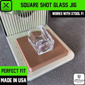 May include: A clear square shot glass sits on a brown base within a metal jig. The jig has the text "SQUARE SHOT GLASS JIG" and "WORKS WITH XTOOL F1". The text "PERFECT FIT" and "MADE IN USA" are also visible.