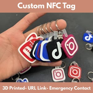 May include: A collection of 3D-printed NFC tags in various colors, including red, blue, black, and pink, each with a social media logo. The tags are attached to keychains and feature the text "Custom NFC Tag" and "3D Printed- URL Link- Emergency Contact".