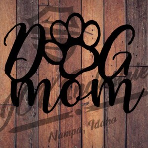 May include: A wooden sign with the words "Dog Mom" in a stylish script font, featuring a paw print design. The sign is set against a rustic, wooden plank background, with the text "Nampa, Idaho" at the bottom.