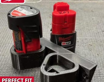 May include: A 3D printed black plastic holder for two Milwaukee M12 batteries. The holder is designed to hold the batteries securely in place. The holder is labeled "PERFECT FIT 3D PRINTED!"