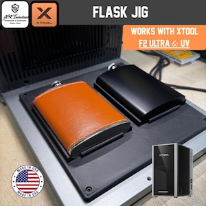 May include: A black flask jig designed for xTool F2 Ultra and UV machines. The device features two recessed slots holding a tan leather-wrapped flask and a matte black metal flask. Text on the unit reads FLASK JIG and WORKS WITH XTOOL F2 ULTRA & UV.