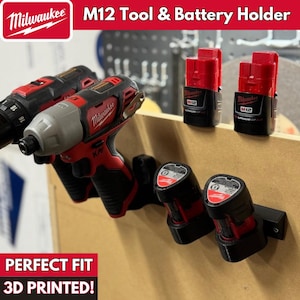 May include: A 3D printed wall mount tool holder for Milwaukee M12 tools and batteries. The holder is black and has two slots for batteries and two slots for tools. The holder is mounted on a light brown surface. The text "PERFECT FIT 3D PRINTED!" is printed below the holder.